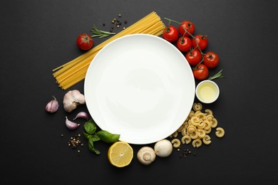 Plate surrounded by different types of pasta, products and peppercorns on black background, flat lay. Space for text