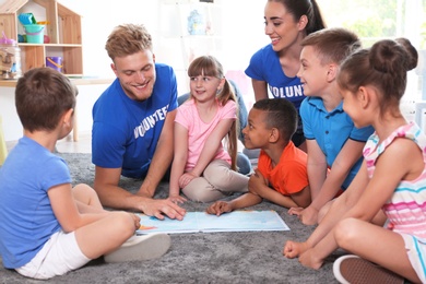 Young volunteers reading book with children on floor indoors