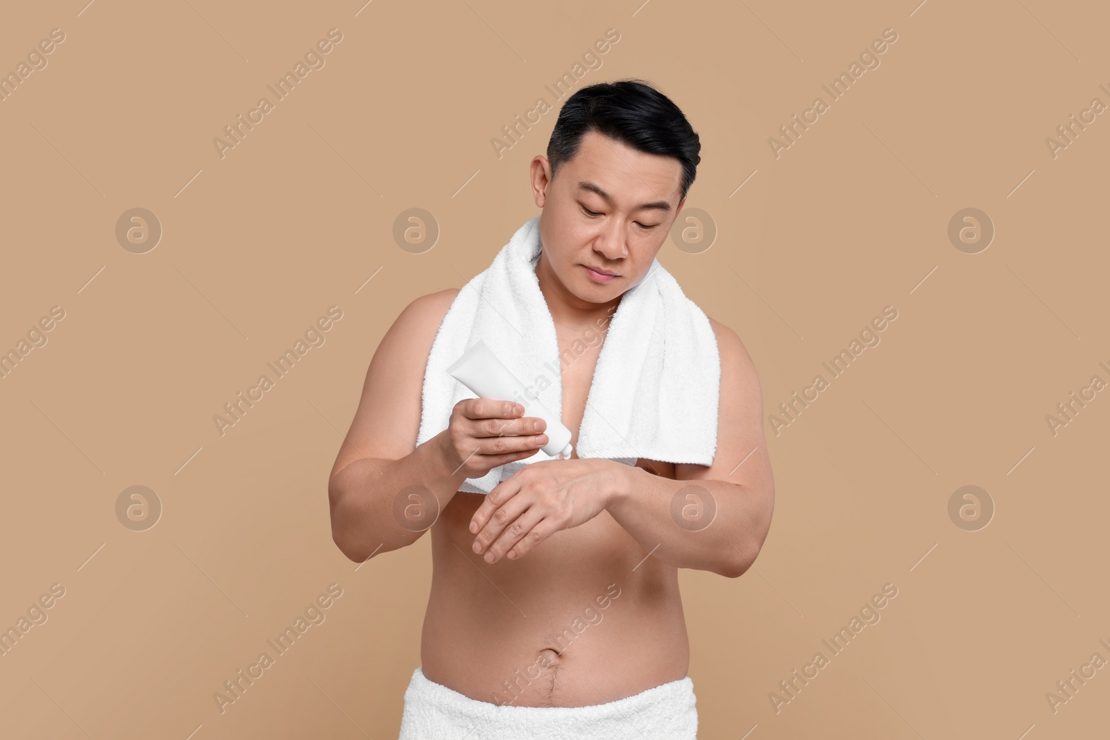 Photo of Man applying body cream from tube onto his hand against light brown background