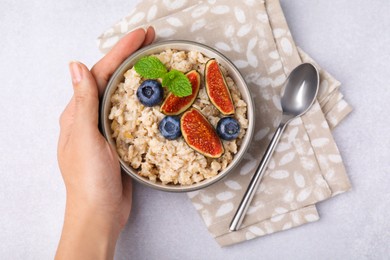 Photo of Woman holding bowl of oatmeal with blueberries, mint and fig pieces at white table, top view