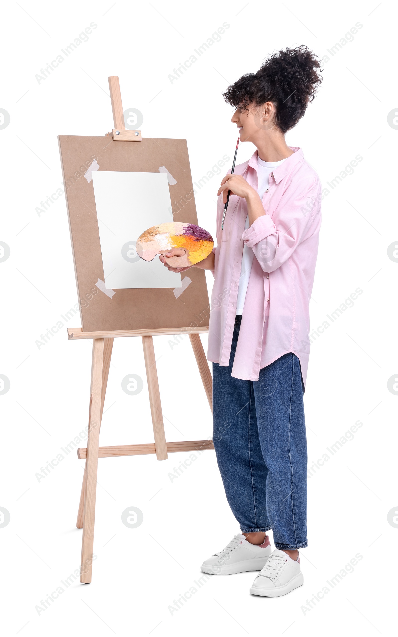 Photo of Young woman holding brush and artist`s palette near easel against white background