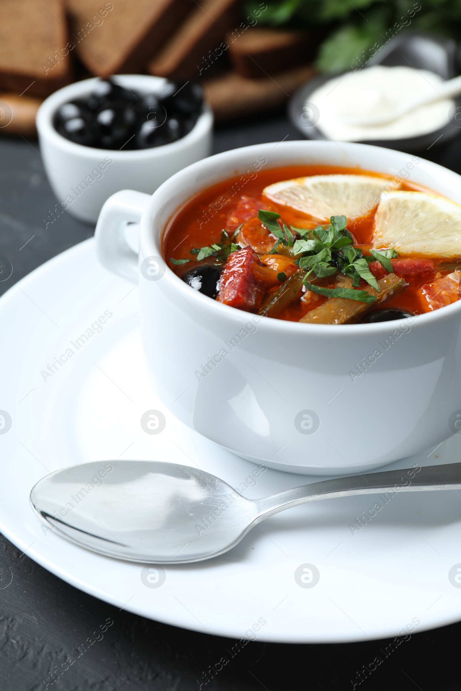 Photo of Meat solyanka soup with sausages, olives, vegetables and spoon on grey table, closeup