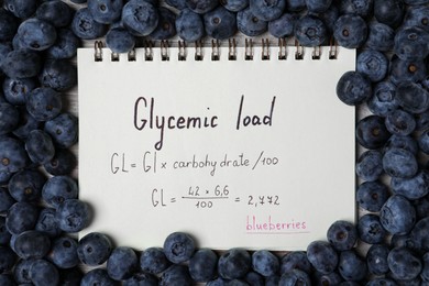 Photo of Notebook with calculated glycemic load for blueberries surrounded by fresh berries on table, top view
