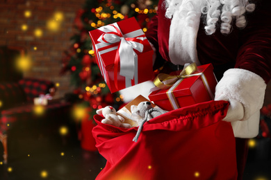 Photo of Santa Claus packing Christmas gifts into bag indoors, closeup