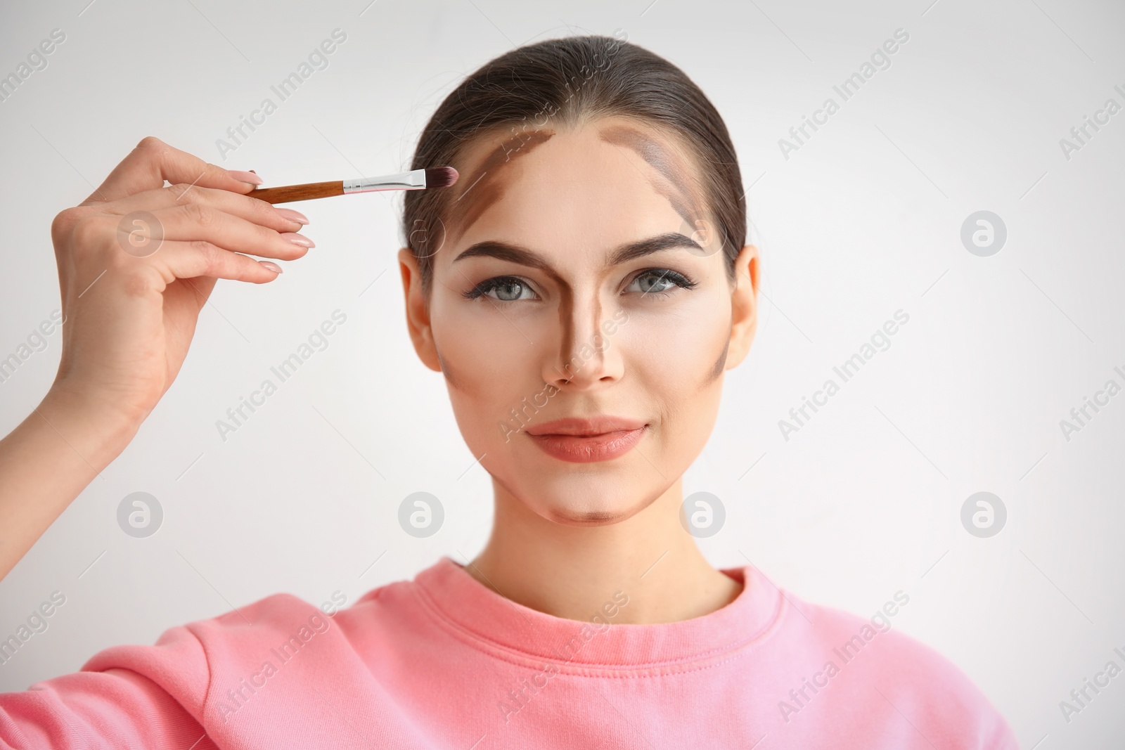 Photo of Young woman applying contouring lines on her face against light background. Professional makeup products