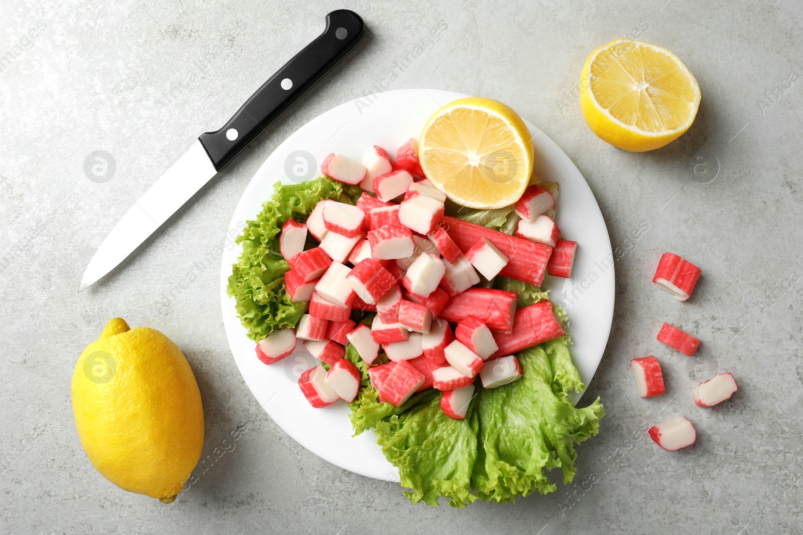 Photo of Cut crab sticks, lettuce, lemons and knife on light table, flat lay