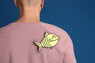 Photo of Man with paper fish on back against blue background, closeup. April fool's day