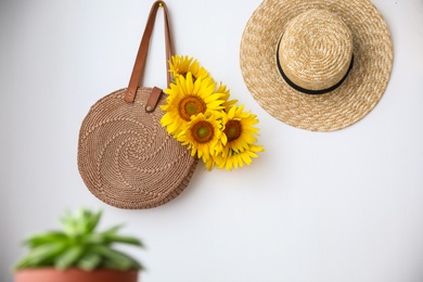 Knitted bag with bouquet of beautiful sunflowers and straw hat on white wall