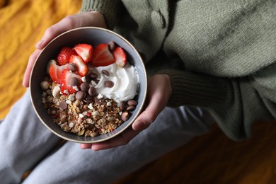 Photo of Woman holding bowl of tasty granola with chocolate chips, strawberries and yogurt indoors, top view