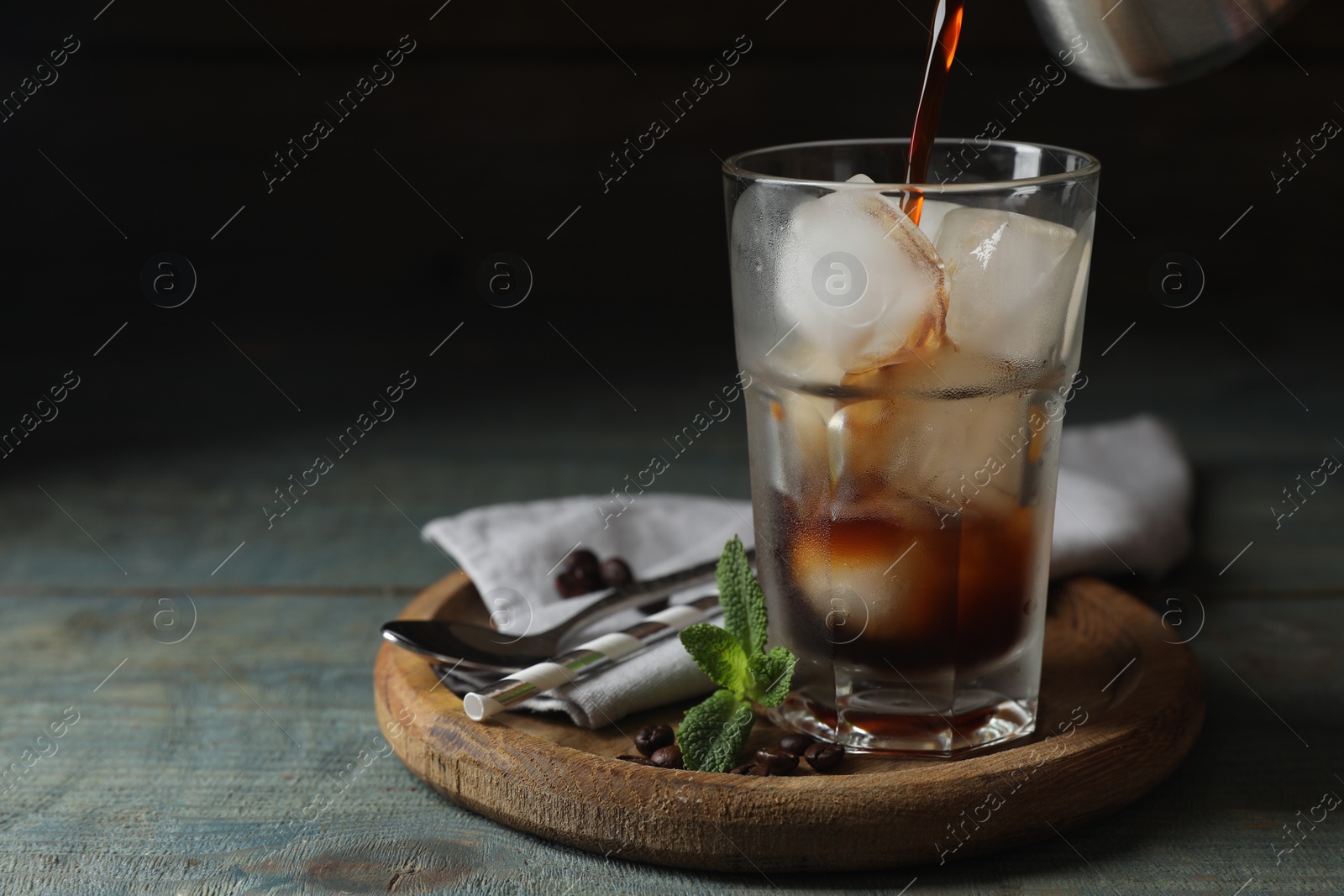 Photo of Pouring coffee into glass with ice cubes, mint and beans on light blue wooden table, space for text