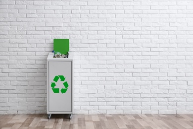Overfilled trash bin with recycling symbol near brick wall indoors. Space for text
