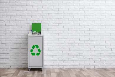 Photo of Overfilled trash bin with recycling symbol near brick wall indoors. Space for text