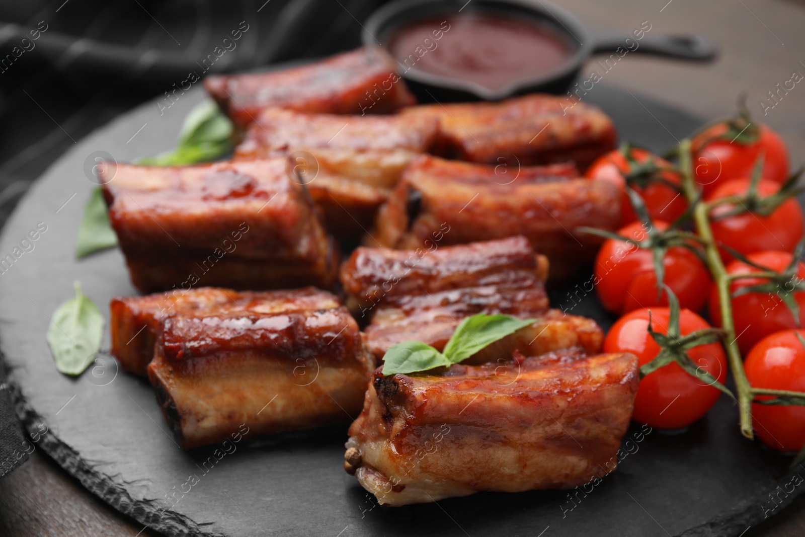 Photo of Tasty roasted pork ribs served with sauce, basil and tomatoes on wooden table, closeup