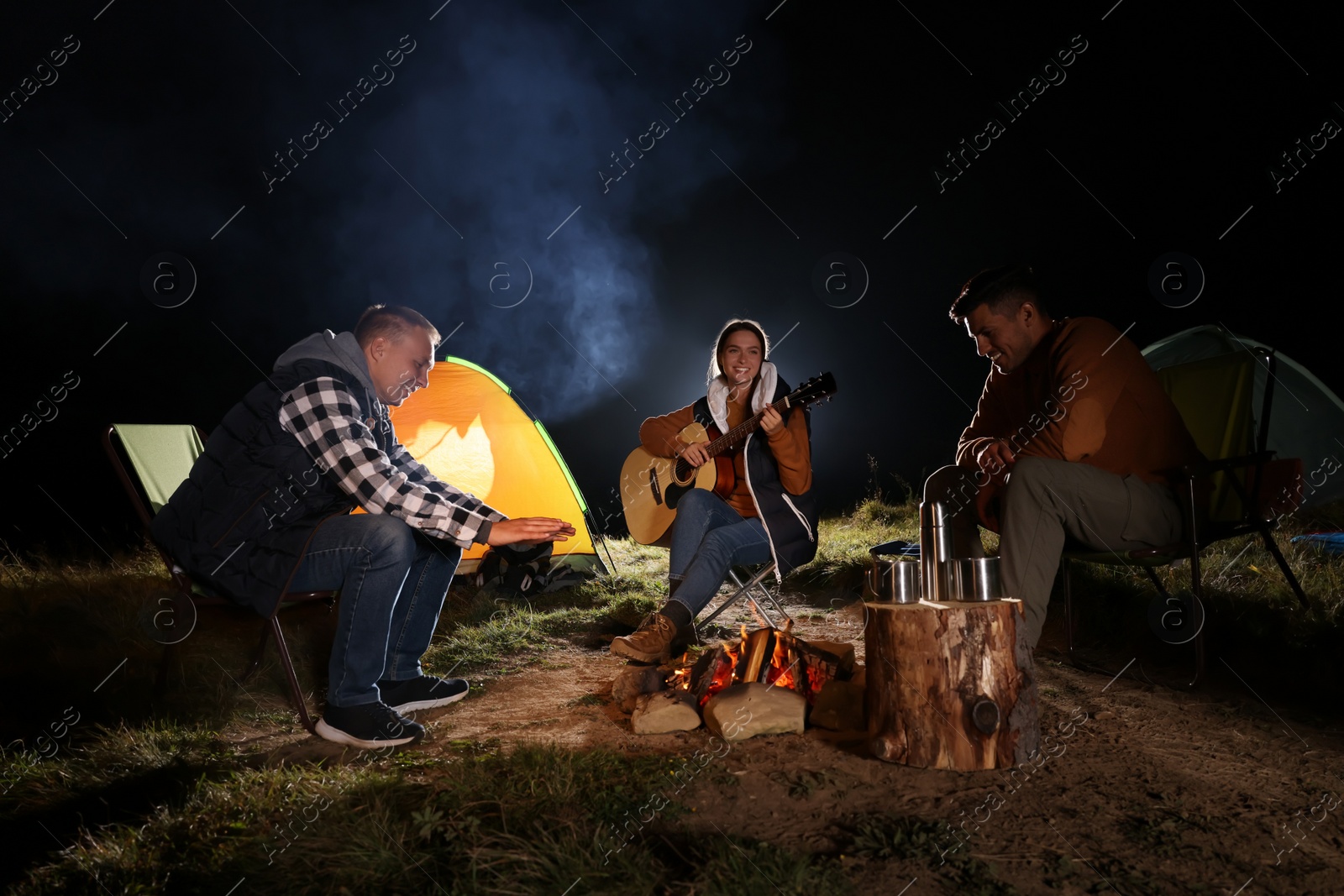 Photo of Group of friends with guitar near bonfire and camping tent outdoors at night