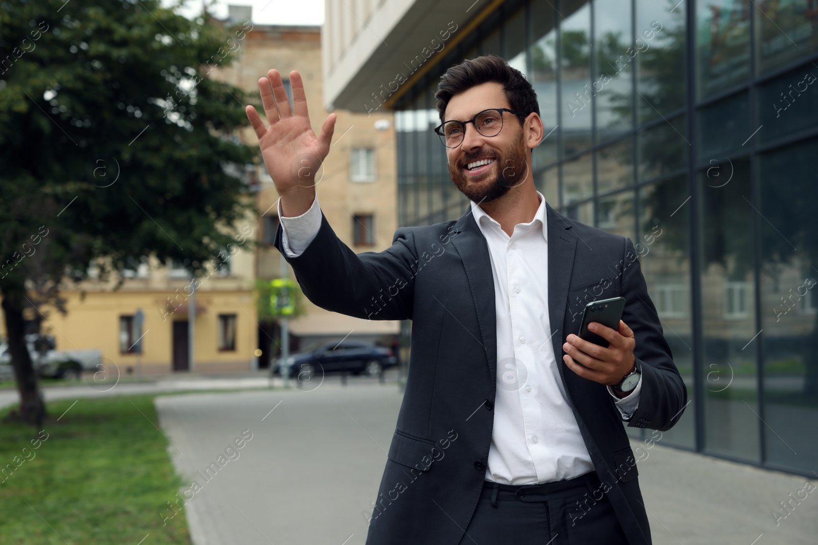 Photo of Handsome businessman with smartphone waving to say hello on city street