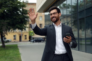 Handsome businessman with smartphone waving to say hello on city street