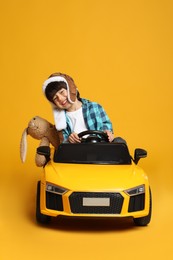 Photo of Cute little boy with toy bunny driving children's car on yellow background