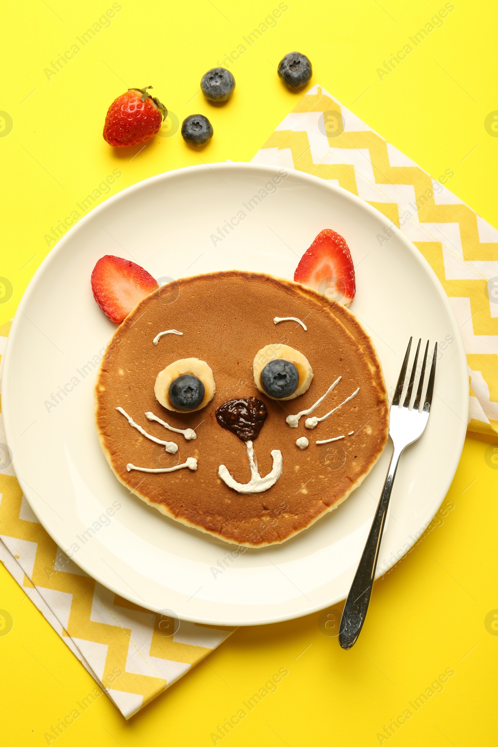 Photo of Creative serving for kids. Plate with cute cat made of pancakes, berries, cream, banana and chocolate paste on yellow background, flat lay