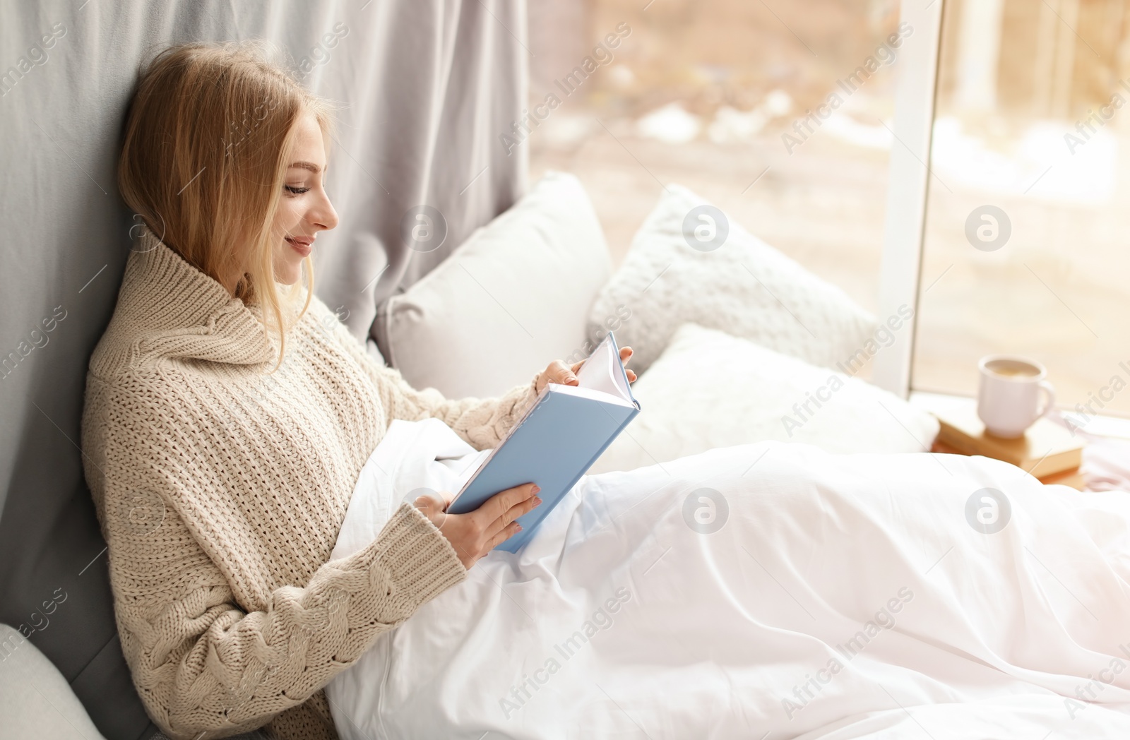 Photo of Beautiful young woman in knitted sweater sitting and reading book near window at home. Winter atmosphere