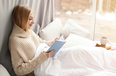 Beautiful young woman in knitted sweater sitting and reading book near window at home. Winter atmosphere