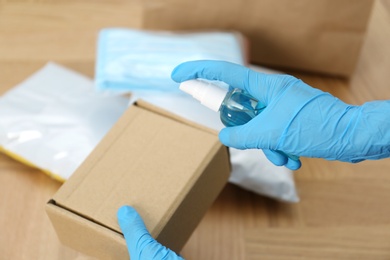 Woman spraying antiseptic onto parcel at wooden table, closeup. Preventive measure during COVID-19 pandemic