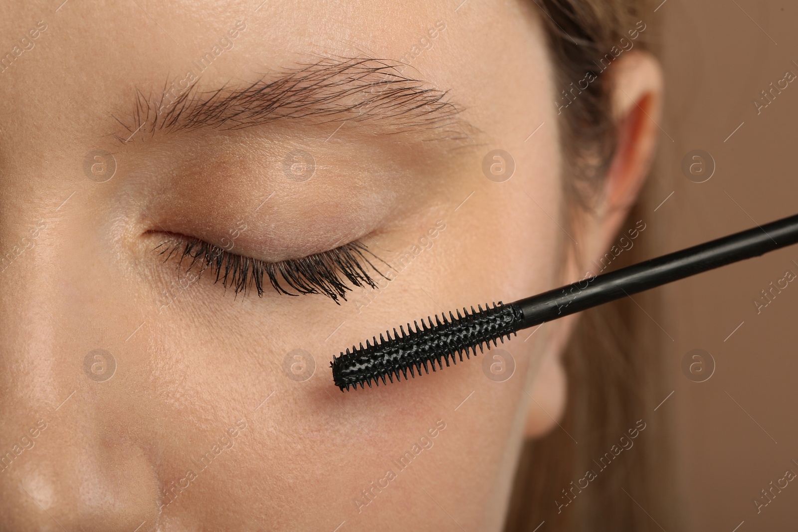 Photo of Woman applying mascara onto eyelashes against light brown background, closeup