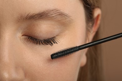 Woman applying mascara onto eyelashes against light brown background, closeup