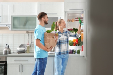 Photo of Happy couple with paper bag full of products standing near refrigerator in kitchen