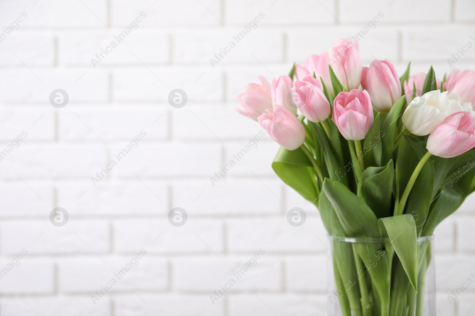 Photo of Beautiful bouquet of tulips in glass vase against white brick wall. Space for text