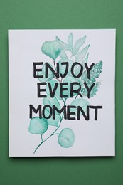 Card with beautiful phrase Enjoy Every Moment on green background, top view