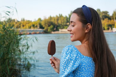 Photo of Beautiful young woman holding ice cream glazed in chocolate near river, space for text