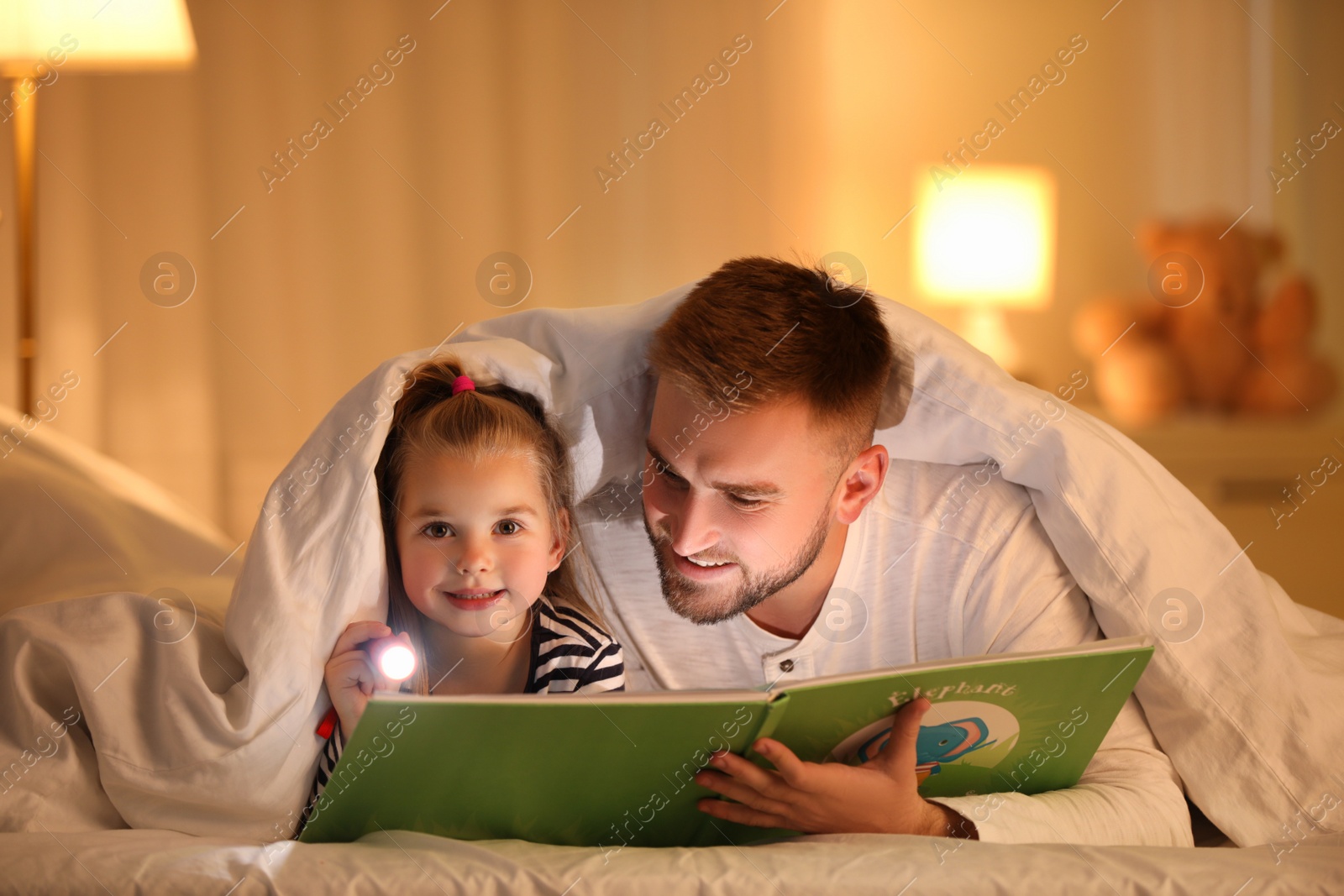 Photo of Father and daughter with flashlight reading book under blanket at home