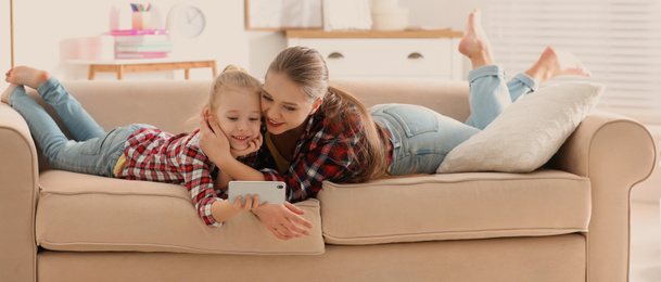 Image of Happy mother and little daughter with smartphone resting on sofa at home. Banner design