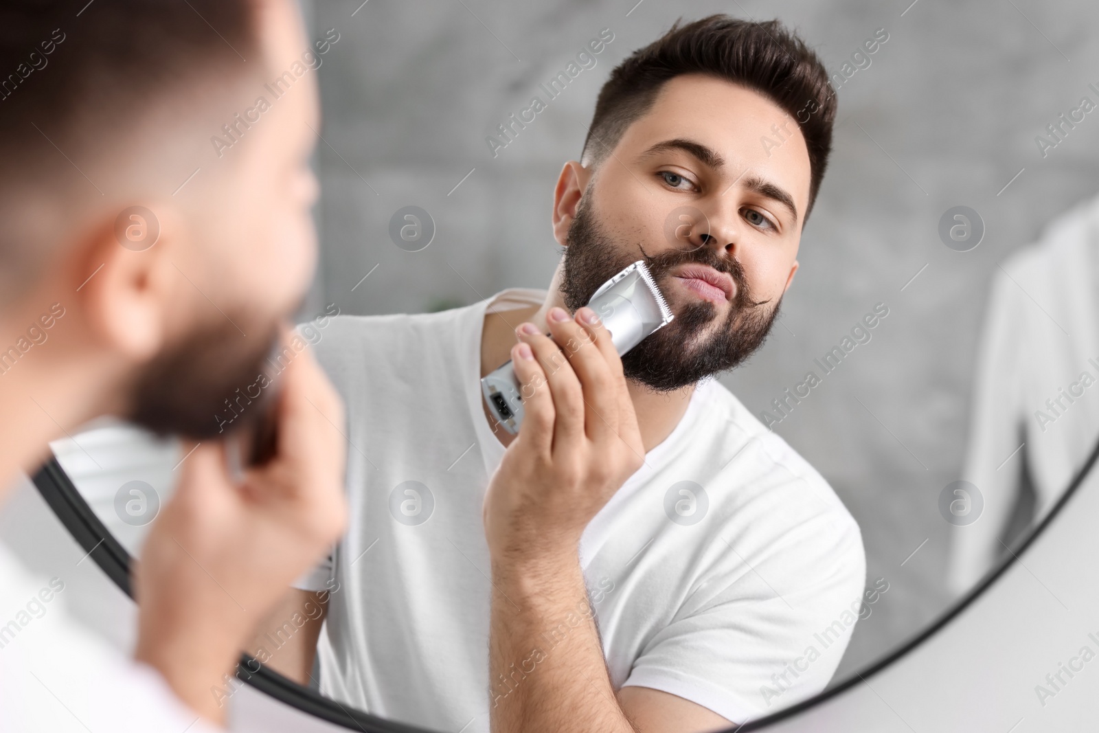 Photo of Handsome young man trimming beard near mirror in bathroom