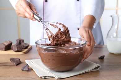 Photo of Professional confectioner whipping chocolate cream with balloon whisk at wooden table, closeup