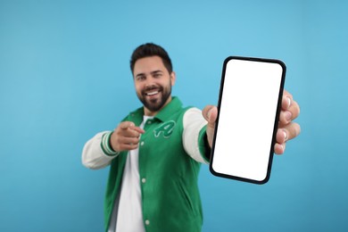 Photo of Young man showing smartphone in hand and pointing at camera on light blue background, selective focus. Mockup for design