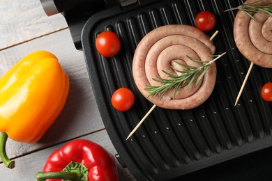 Photo of Electric grill with homemade sausages, rosemary and vegetables on rustic wooden table, flat lay