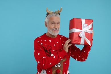 Photo of Senior man in Christmas sweater and reindeer headband holding gift on light blue background
