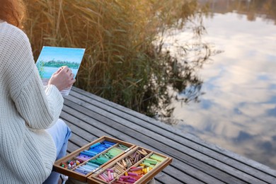 Photo of Woman drawing with soft pastels on wooden pier near river, closeup