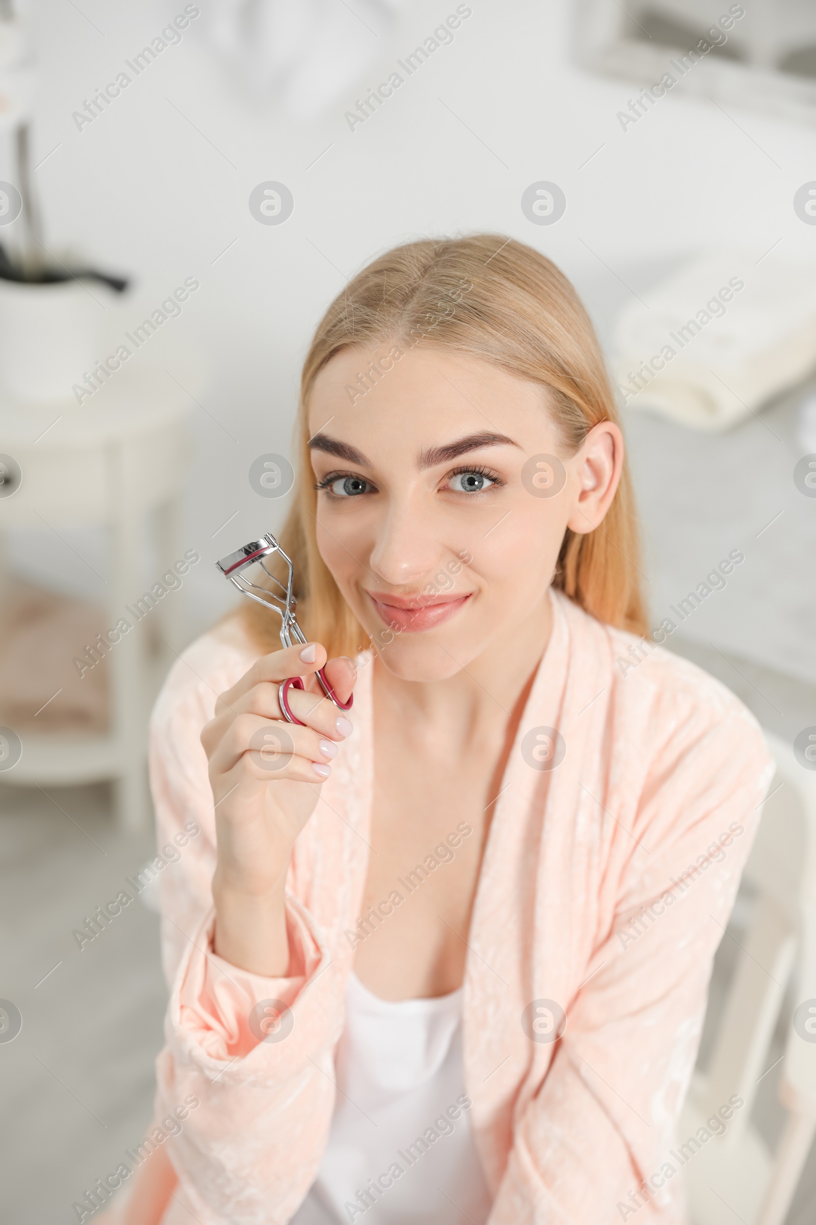 Photo of Attractive young woman curling her eyelashes indoors