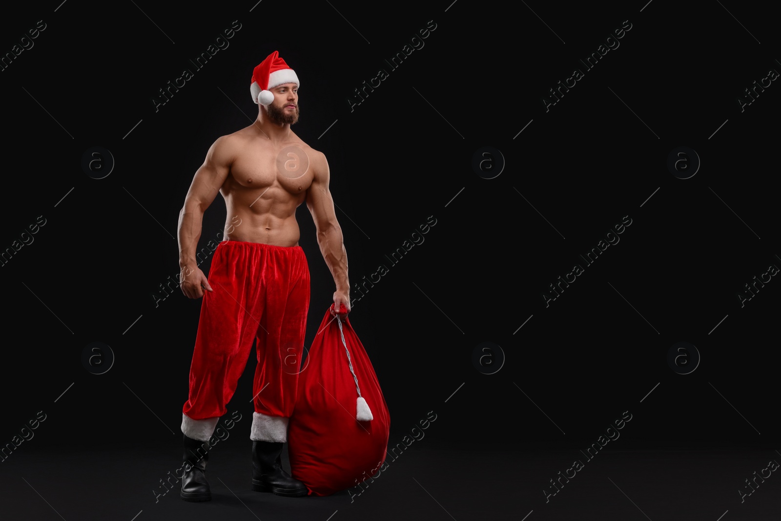 Photo of Muscular young man in Santa hat holding bag with presents on black background, space for text