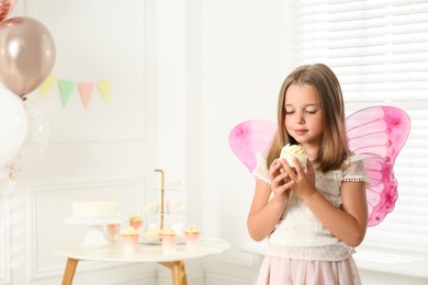 Photo of Cute little girl wearing fairy costume with delicious cupcake in decorated room. Space for text