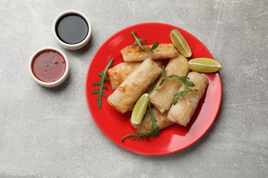 Photo of Tasty fried spring rolls, arugula, lime and sauces on grey textured table, flat lay