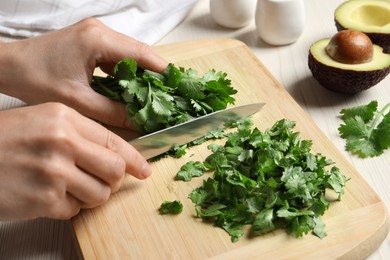 Photo of Woman cutting fresh green cilantro at white wooden table, closeup