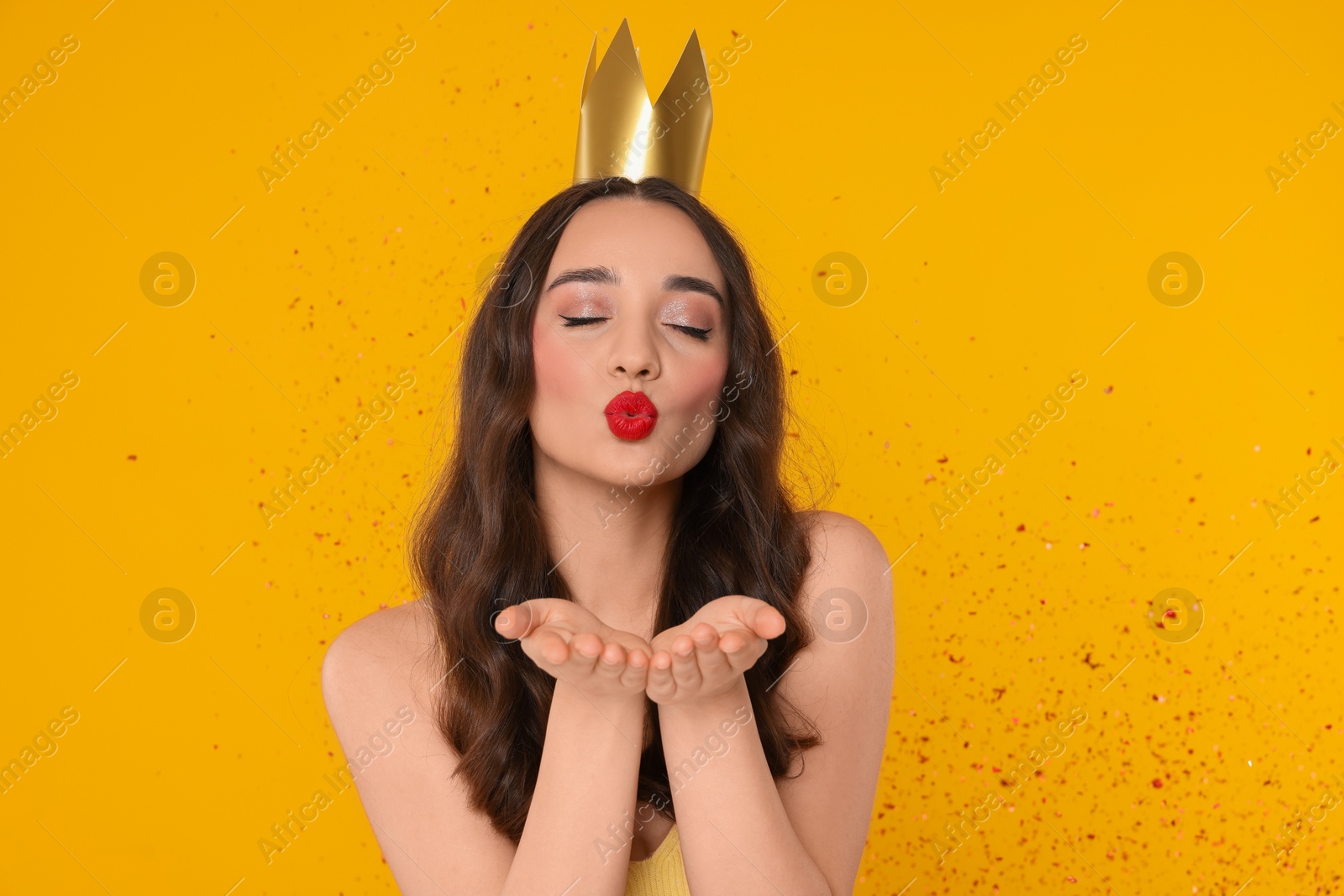 Photo of Beautiful young woman with princess crown blowing kiss under falling confetti on yellow background