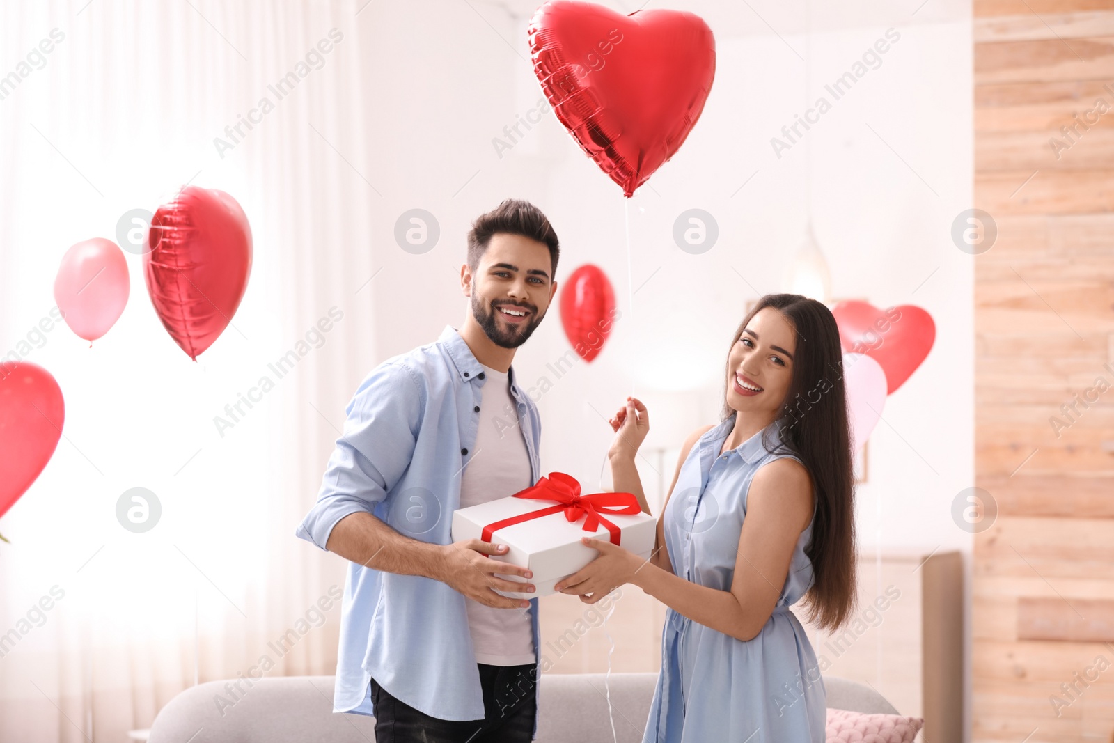 Photo of Young man presenting gift to his girlfriend in living room decorated with heart shaped balloons. Valentine's day celebration