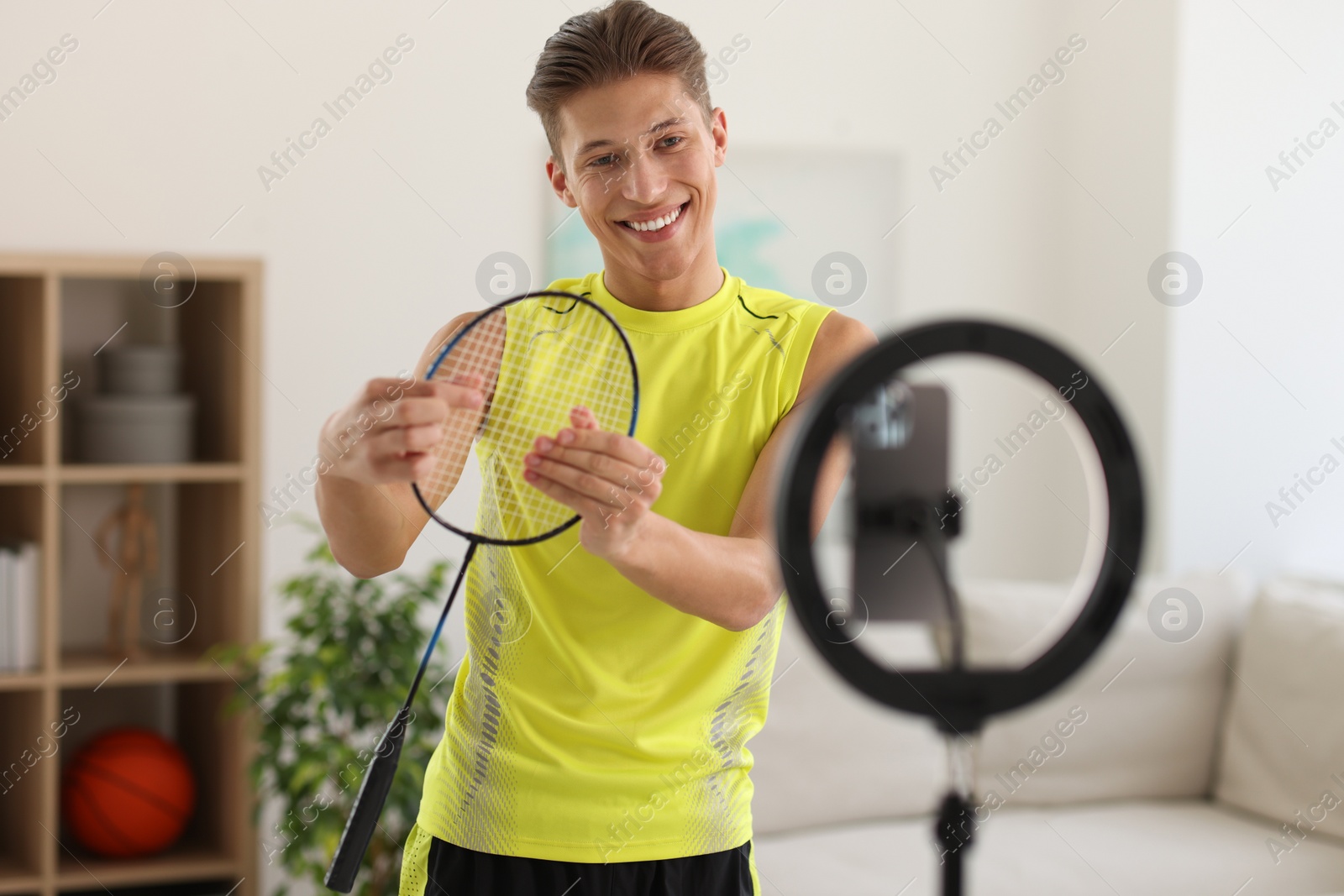 Photo of Smiling sports blogger holding badminton racket while streaming online fitness lesson with smartphone at home