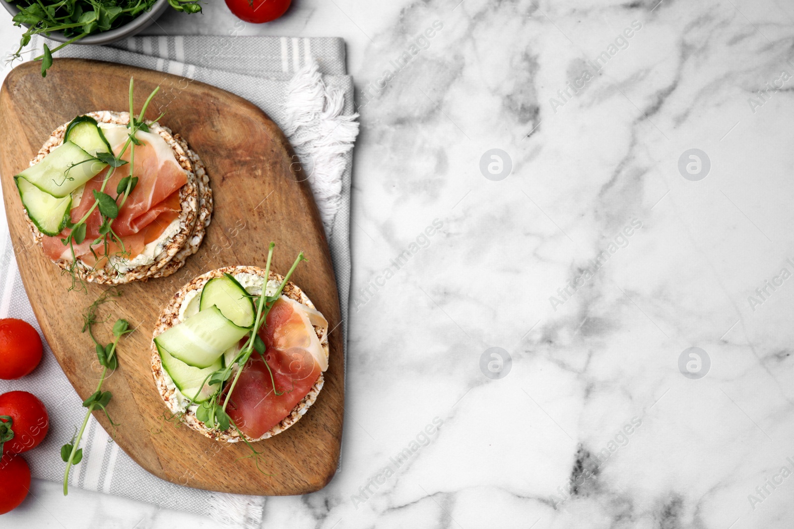 Photo of Crunchy buckwheat cakes with cream cheese, prosciutto and cucumber slices on white marble table, flat lay. Space for text