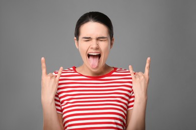 Photo of Happy young woman showing her tongue and making rock gesture on grey background