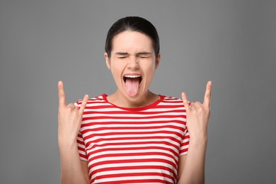 Happy young woman showing her tongue and making rock gesture on grey background